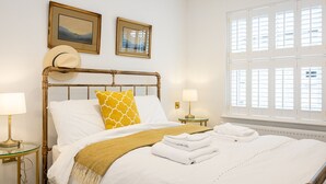 A neatly made bed with white linens and a yellow patterned pillow, a bedside table with a lamp, and a window with blinds.