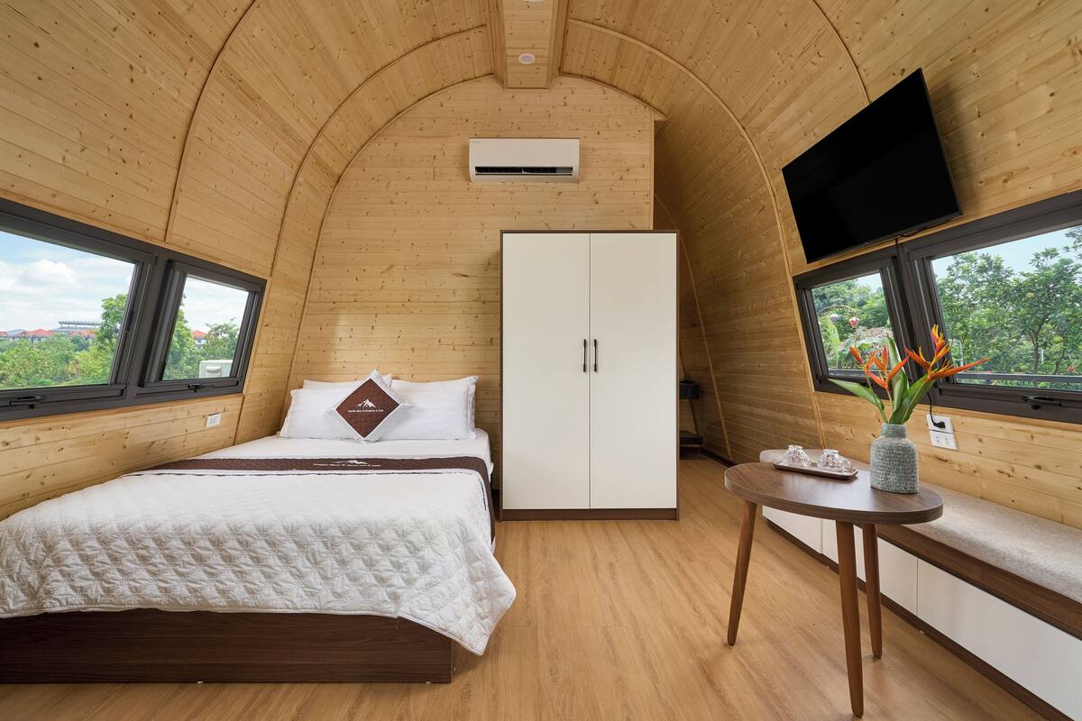 A bedroom with a bed, a wardrobe, a TV, and a window with a view of greenery.