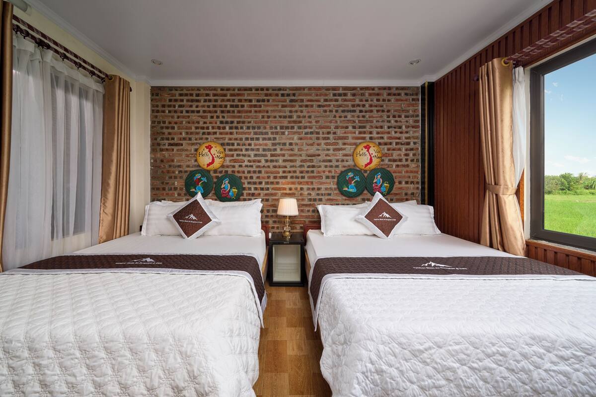 A hotel room with two beds, a brick wall, a window with curtains, and a nightstand with a lamp.