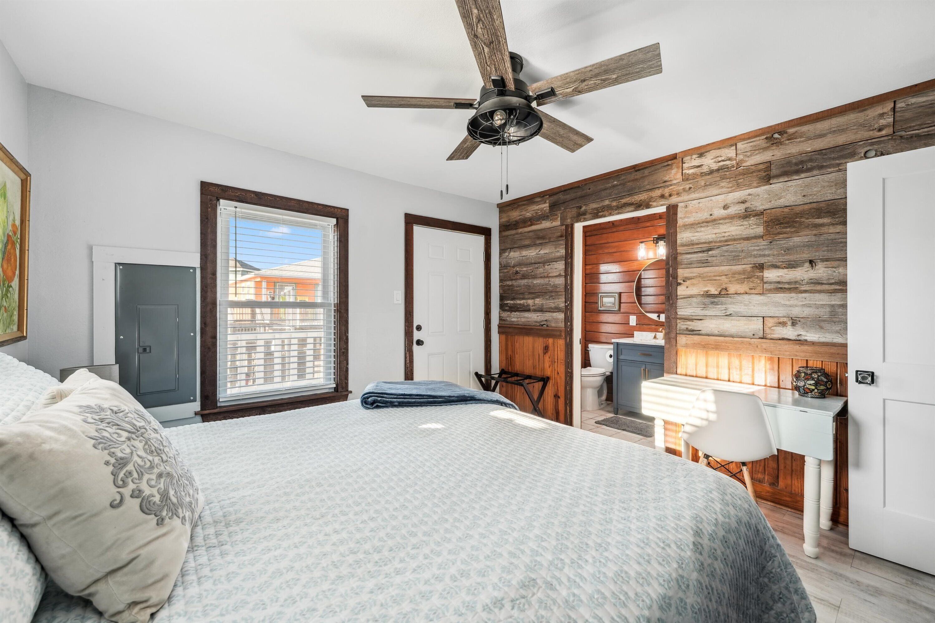 A bedroom with a bed, a ceiling fan, a wooden wall panel, a window, and a door.