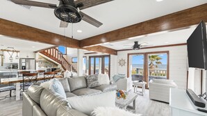 A spacious living area with a gray leather sofa, a white coffee table, and a ceiling fan. A kitchen is visible in the background with a dining area and a staircase leading to a balcony. - Stunning Beach Front Paradise (Surfside Beach)