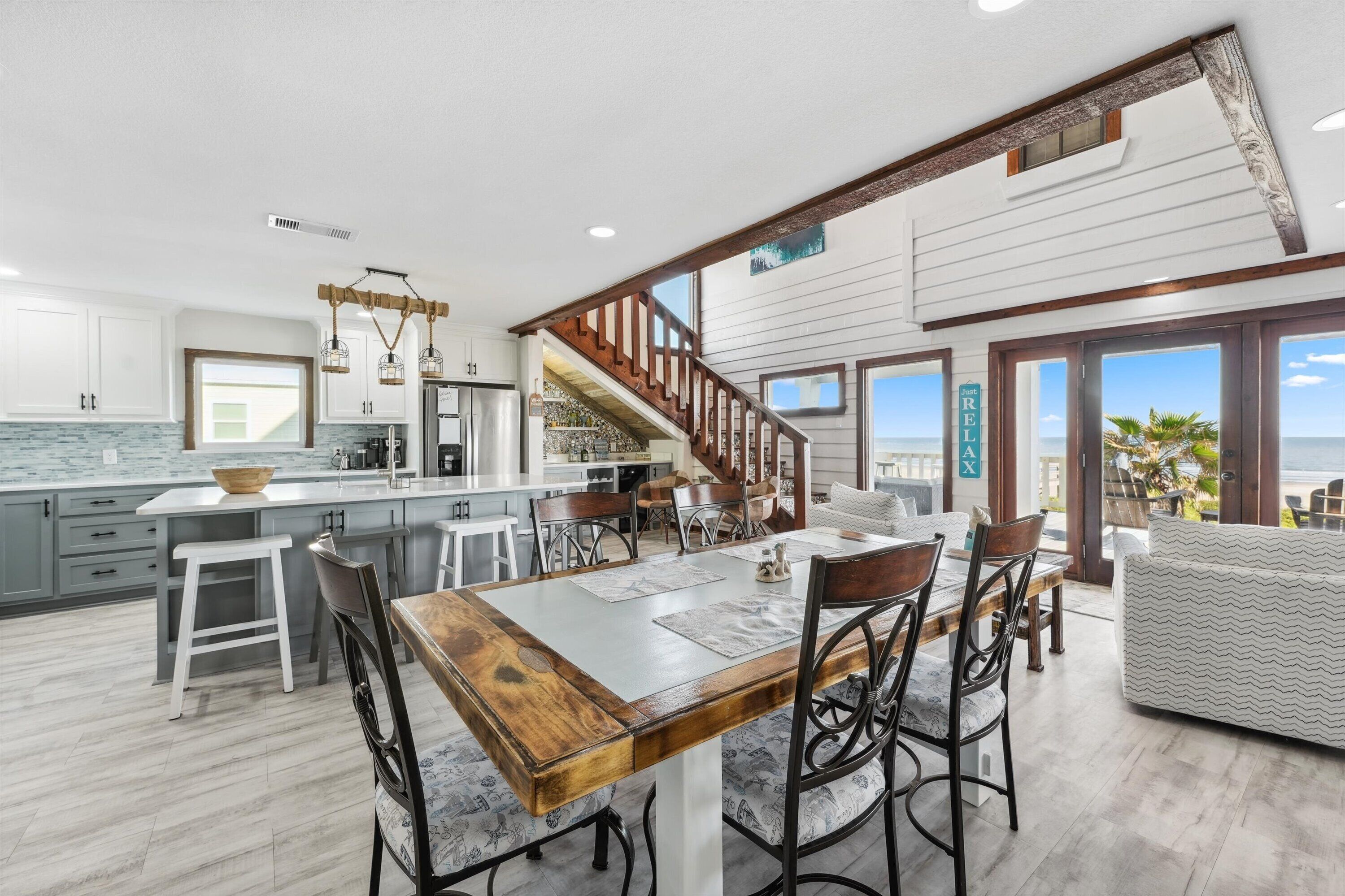 Une cuisine spacieuse dotée d’un grand îlot central, d’une table à manger en bois et de tabourets de bar modernes. Un escalier mène à un balcon offrant une vue sur l’océan.