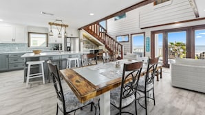A spacious kitchen with a large island, wooden dining table, and modern bar stools. A staircase leads to a balcony with a view of the ocean. - Stunning Beach Front Paradise (Surfside Beach)