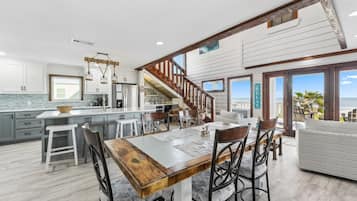 Une cuisine spacieuse dotée d’un grand îlot central, d’une table à manger en bois et de tabourets de bar modernes. Un escalier mène à un balcon offrant une vue sur l’océan.