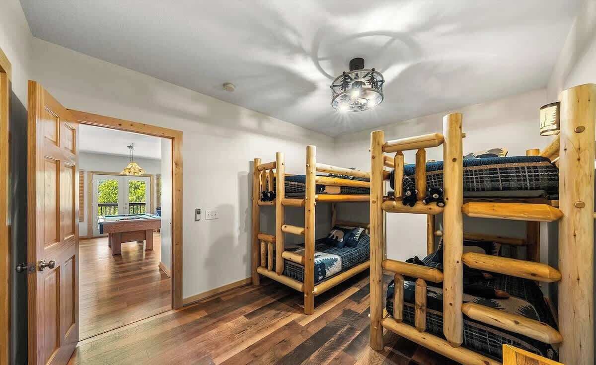 A room with bunk beds, wooden flooring, a ceiling light, and a door leading to another room with a pool table.