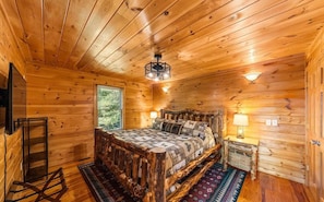 Quarto de cabana de madeira com cama, mesa de cabeceira, abajur, janela e ventilador de teto.