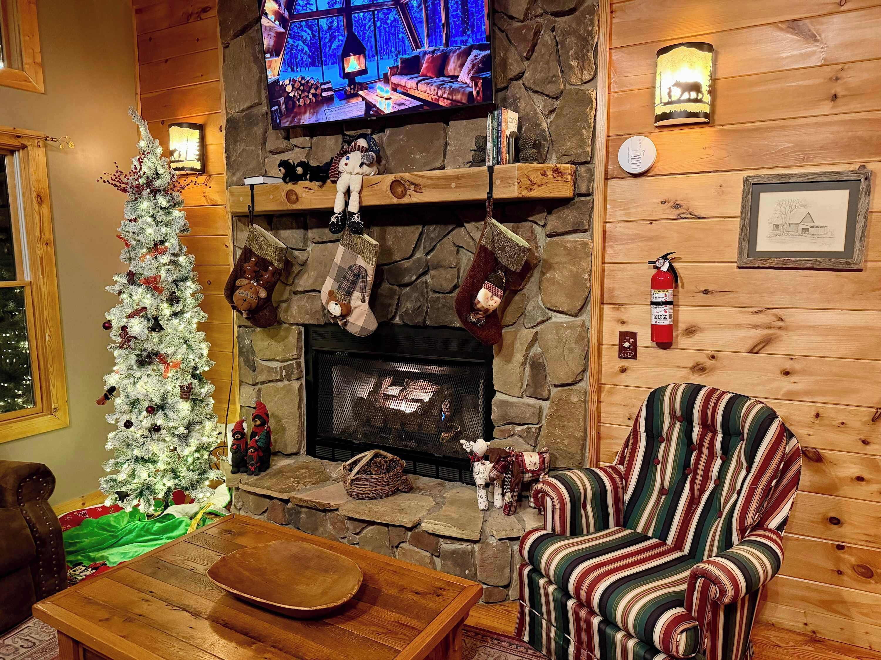 Un salon chaleureux avec une cheminée en pierre, un sapin de Noël et un téléviseur diffusant une scène hivernale enneigée.