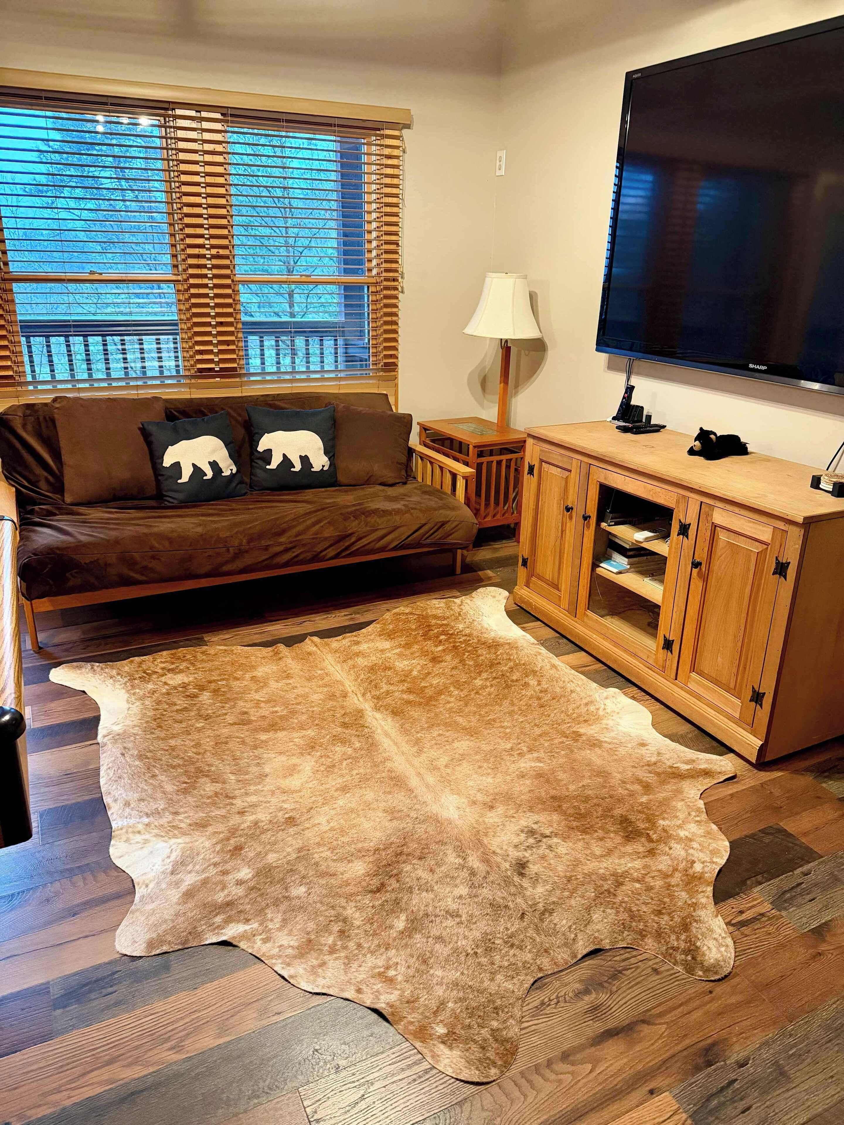Un salon chaleureux avec un canapé en cuir marron, deux coussins en forme d’ours, un meuble en bois, un grand téléviseur à écran plat et un grand tapis marron.