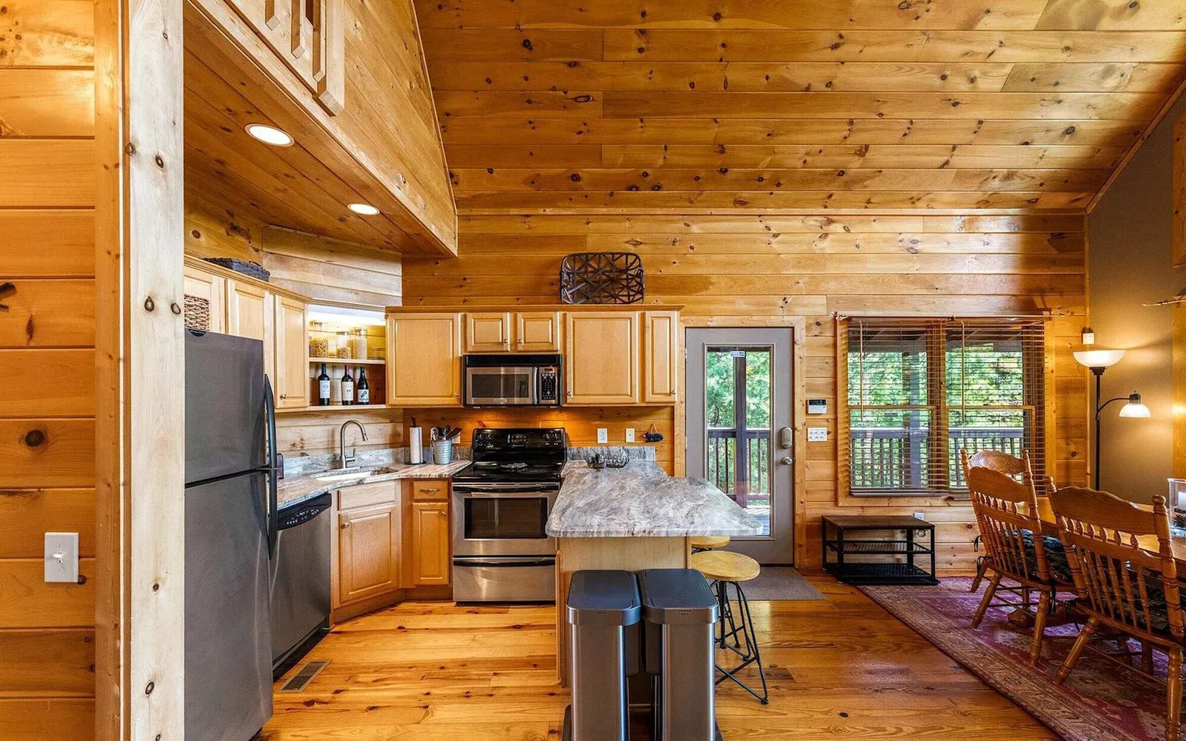 A cabin-style kitchen with wooden walls, a refrigerator, stove, microwave, and a dining area with a table and chairs.