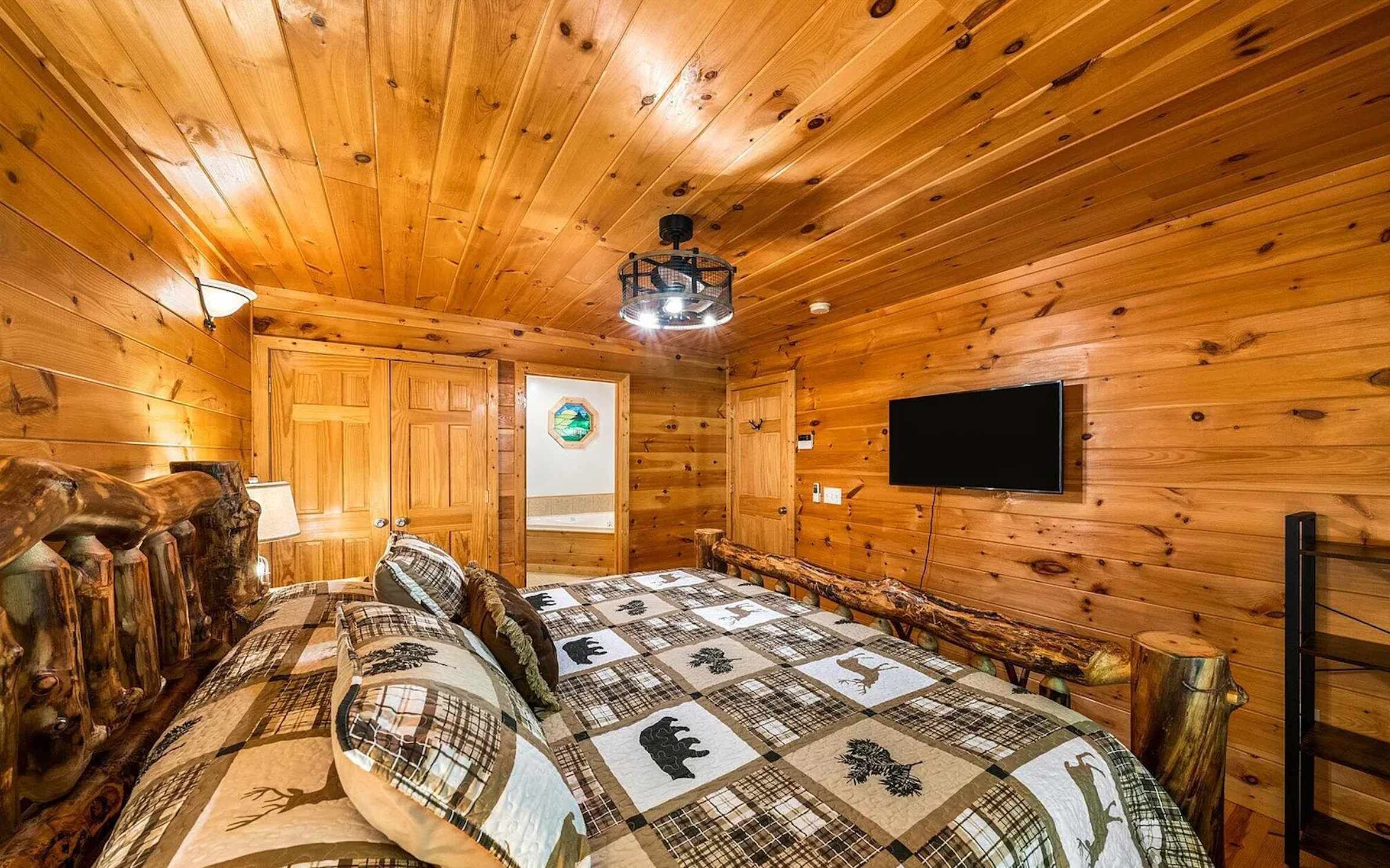 Une chambre confortable, aménagée dans le style d’un chalet, avec un plafond en bois, un lit recouvert d’une courtepointe à motifs, un téléviseur fixé au mur et un banc en bois rustique.