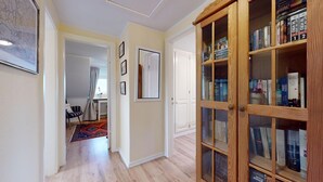 Un pasillo con una estantería de madera llena de libros, un mapa enmarcado en la pared y una puerta que da a otra habitación.