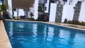 En swimmingpool med klart blåt vand, omgivet af en flisbelagt terrasse og en bygning med altaner i baggrunden.