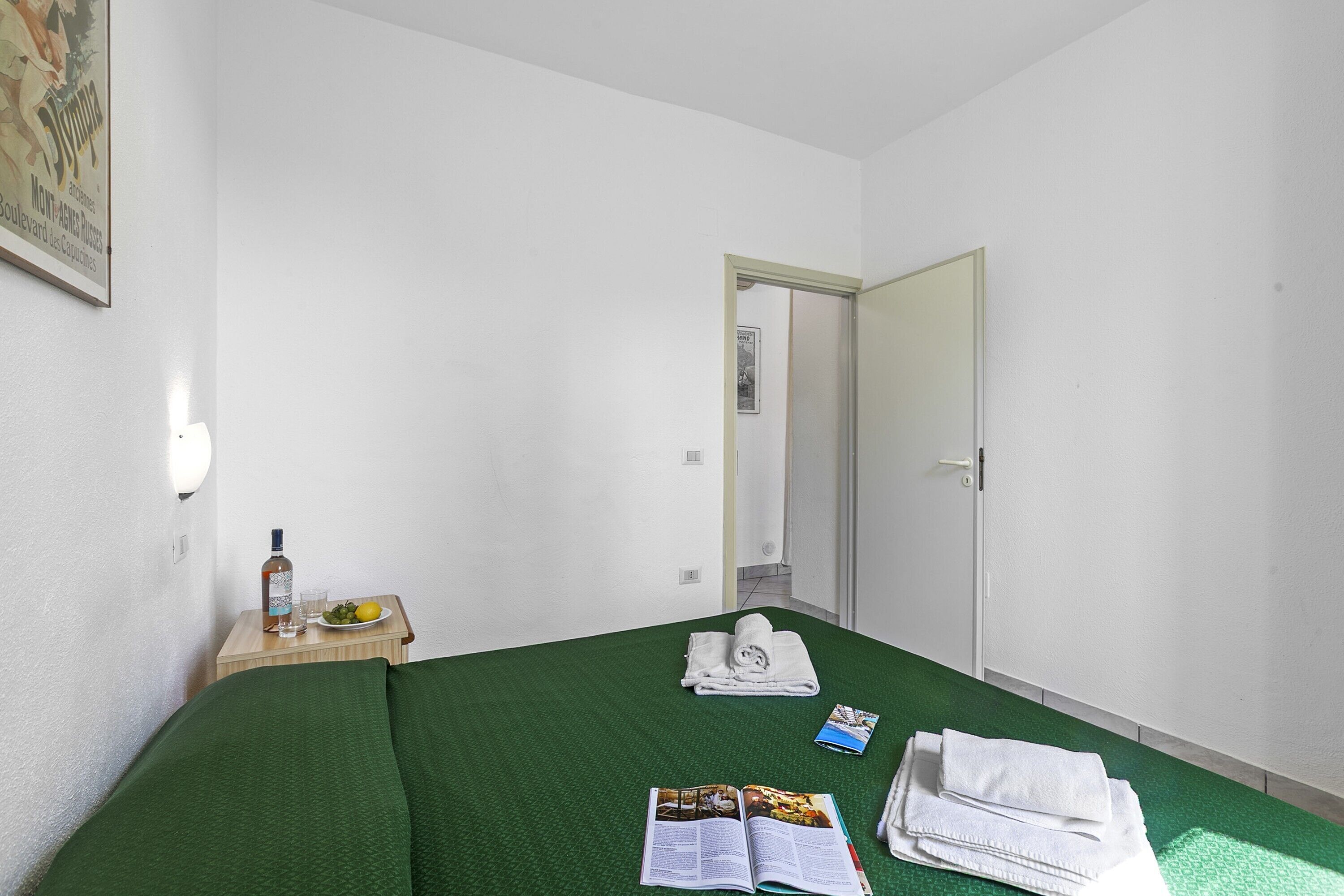 Ett hotellrum med ett grönt sängöverkast, ett litet bord med en flaska, glas och en tallrik med frukt, en dörr och en inramad tavla på väggen.