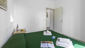 Habitación de hotel con una colcha verde, una pequeña mesa con una botella, vasos y un plato de fruta, una puerta y un cuadro en la pared.