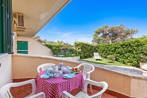 A balcony with a checkered table set for dining, overlooking a garden with trees and a hedge.