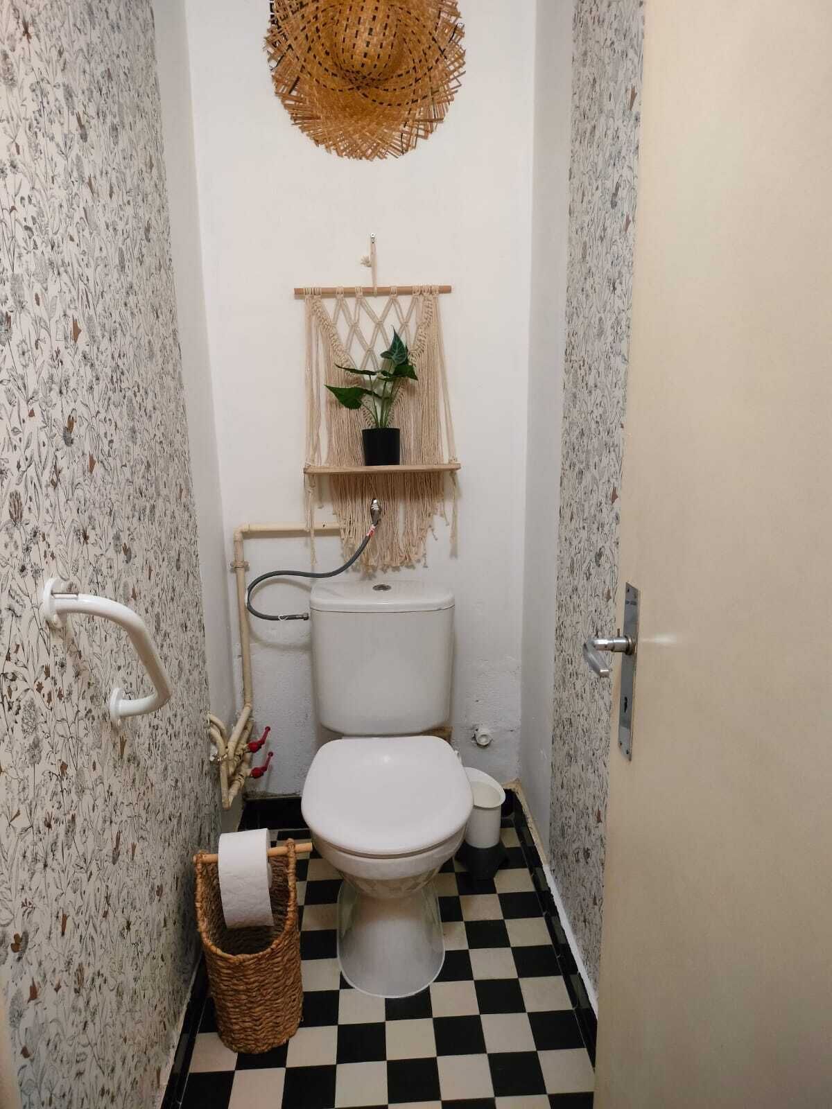 A small bathroom with a macramé shelf holding a potted plant, a toilet, and a checkered floor.