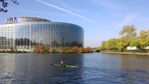 Un bâtiment moderne en verre, situé au bord d’une rivière paisible, avec un bateau à rames au premier plan.