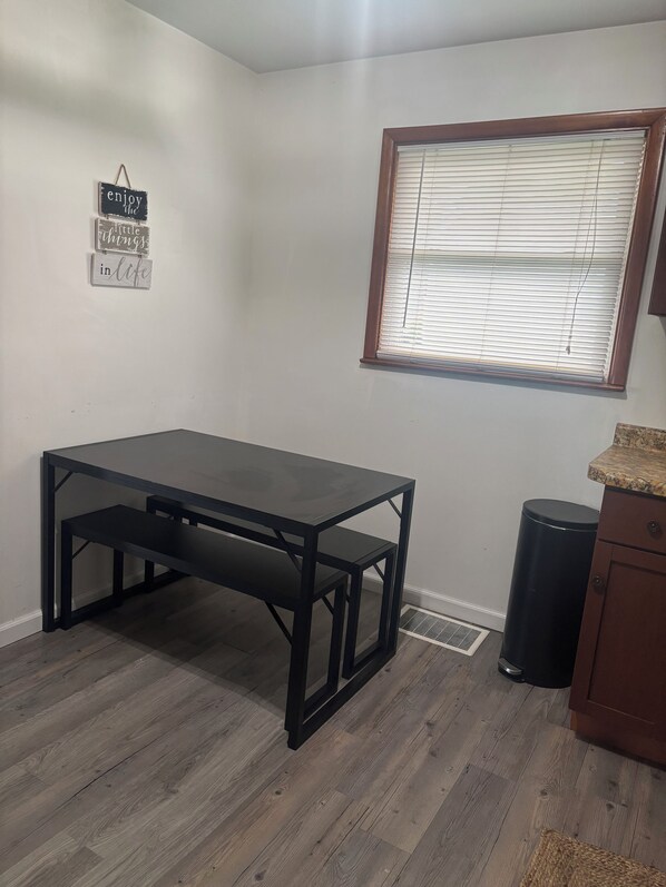 Une pièce avec une table en bois foncé, une fenêtre avec des stores, une petite poubelle et un meuble.