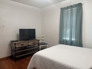 A bedroom with a bed, a TV on a wooden dresser, a chair, and a window with curtains.
