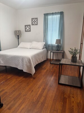 A bedroom with a bed, a nightstand, a lamp, a window with curtains, and a wall with decorative items.