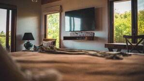 A bedroom with a bed, a lamp, a TV mounted on the wall, and a window with a view of greenery.