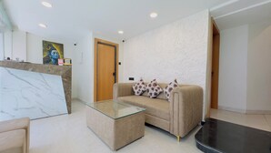 A modern living room with a beige sofa, a glass coffee table, and a marble accent wall. - Hotel Sharda Inn (Ujjain)