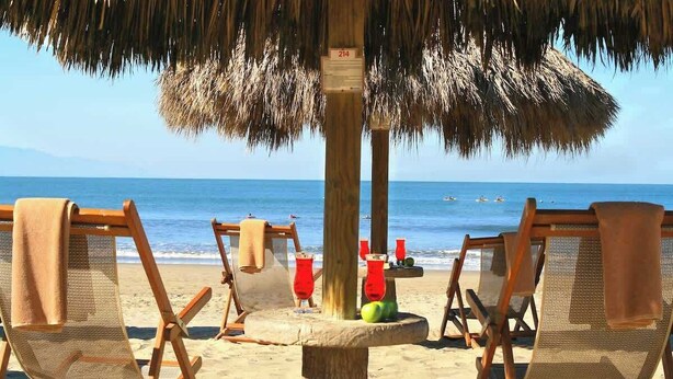 A beach scene with wooden lounge chairs under a thatched umbrella, a wooden post with a sign, and the ocean in the background.