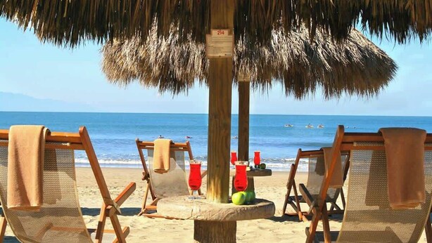 Una playa con sillas de madera para tomar el sol bajo un toldo de paja, un poste de madera con un letrero y el océano al fondo.