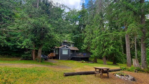 Une cabane entourée d’une forêt dense, avec une terrasse et une table de pique-nique au premier plan.