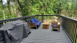 Une terrasse en bois, agréée de mobilier d’extérieur, notamment une table et des chaises abritées, le tout entouré d’une végétation luxuriante.
