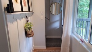 Un couloir avec un tapis à motifs, des œuvres d’art encadrées accrochées au mur, une suspension décorative contenant des plantes vertes et une fenêtre avec des rideaux.