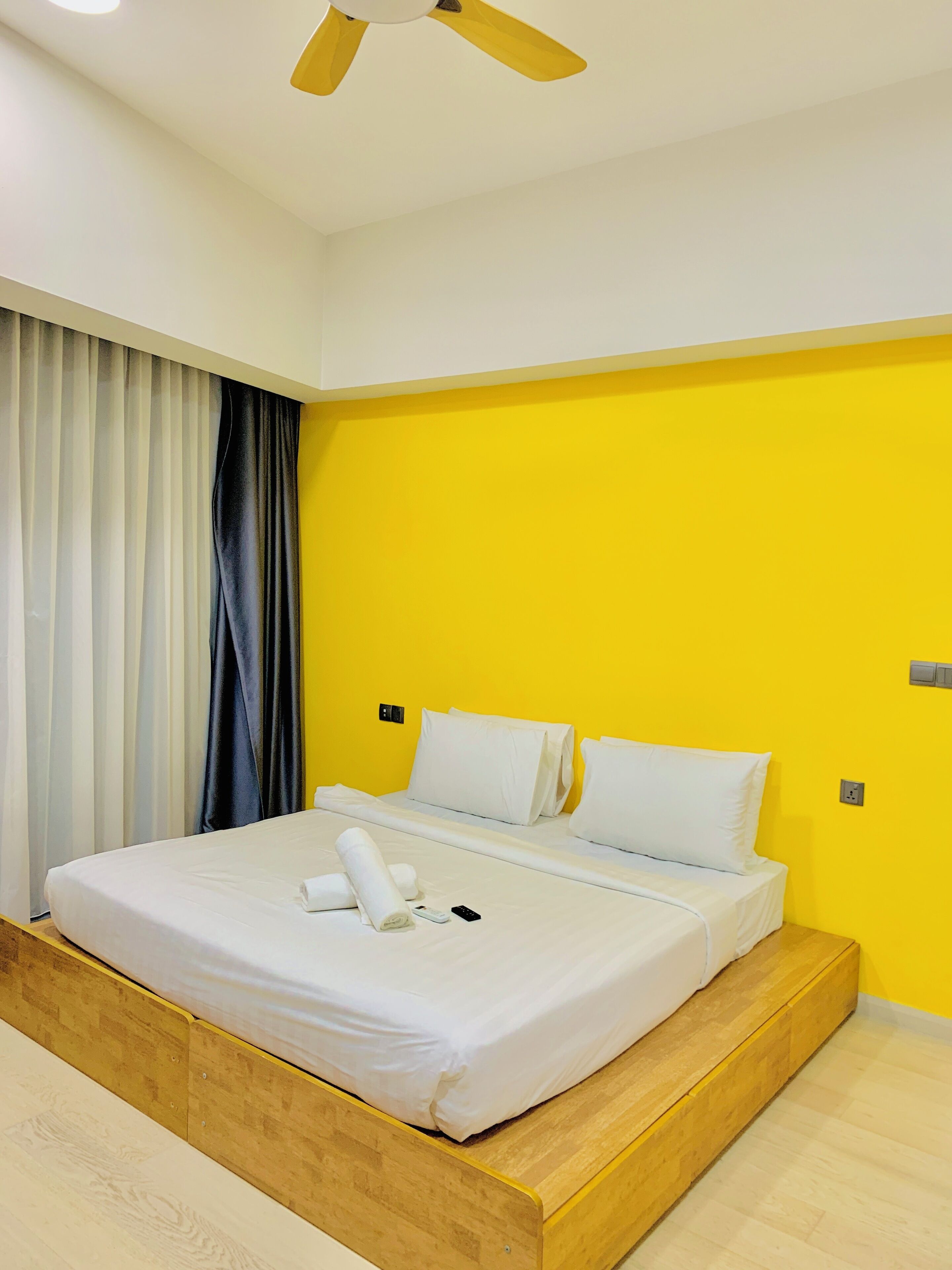 Quarto de hotel moderno com cama, plataforma de madeira, cortinas e ventoinha no teto.