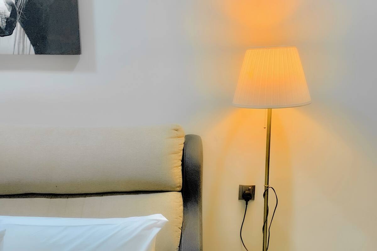 A bed with white linens, a black headboard, and a white pillow. A floor lamp with a yellow base and a white lampshade is on the floor next to the bed.