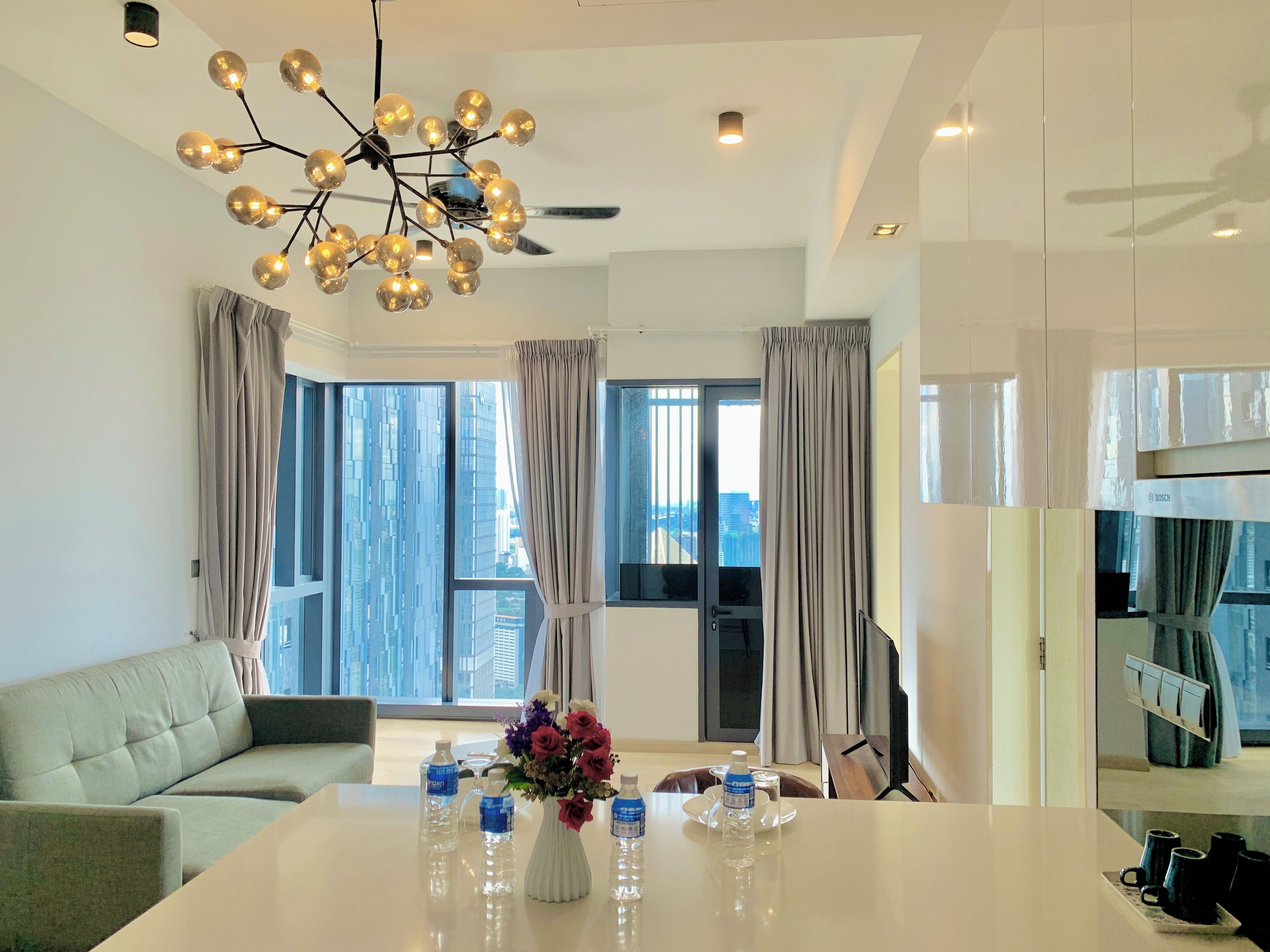 Una habitación de hotel moderna con un ventanal grande, un sofá, una mesa de comedor con botellas de agua y un jarrón de flores, y una lámpara en el techo.