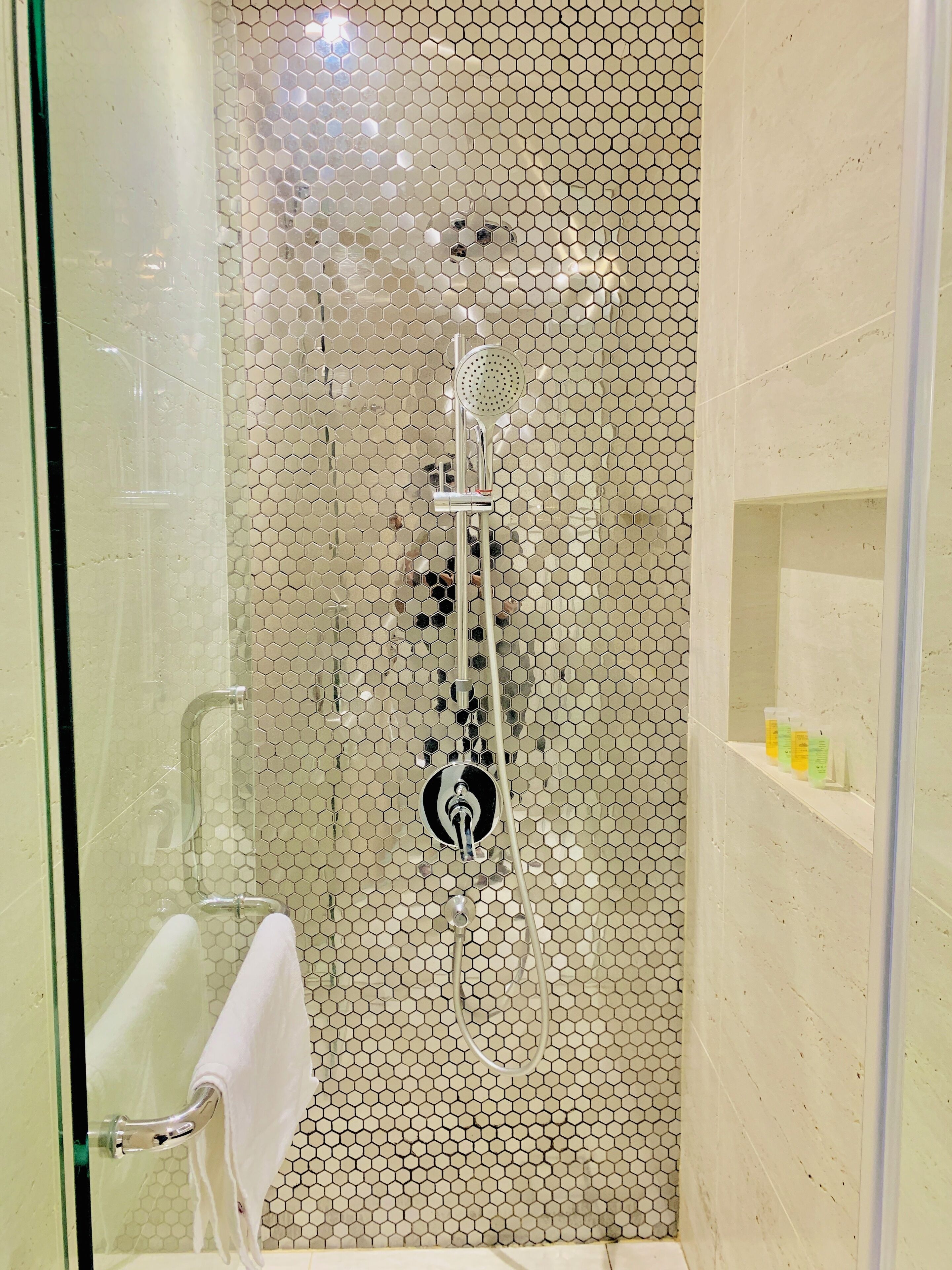 Ducha con cerramiento de vidrio, patrón de azulejos hexagonales, cabezal de ducha de mano y toallero.
