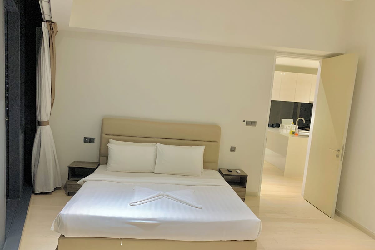 A neatly made bed in a hotel room with a headboard, a bedside table with a remote control, and a visible bathroom in the background.