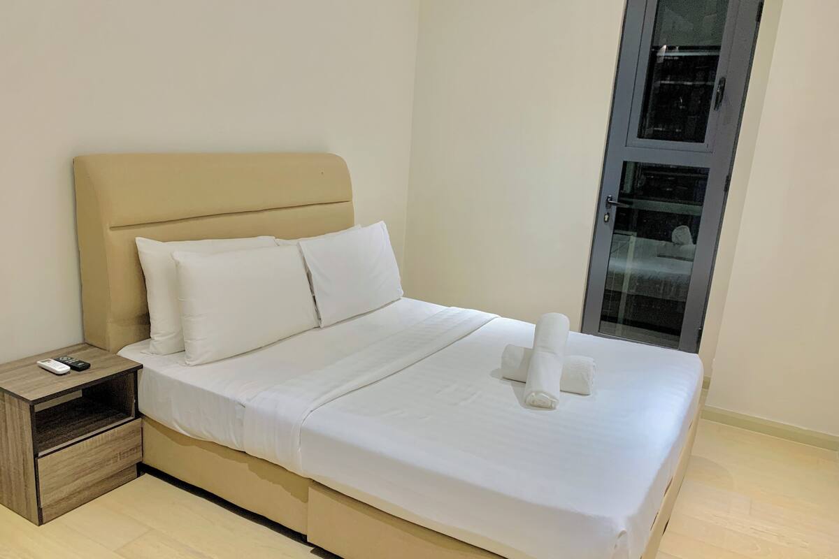 A neatly made bed with white linens, a wooden nightstand with a remote control, and a glass door to the right.
