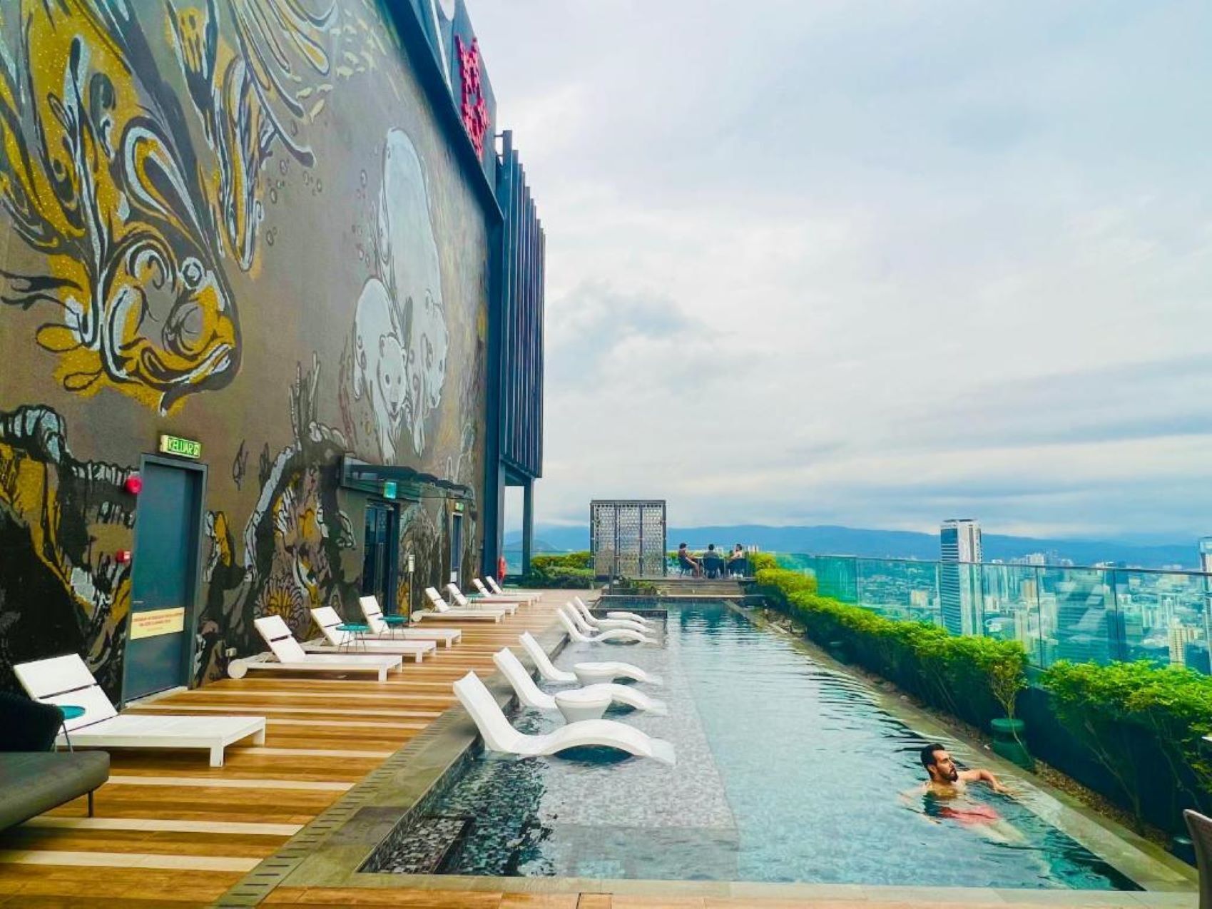Ein Pool mit überlaufendem Rand auf dem Dach, ausgestattet mit Liegestühlen und einer Wandmalerei an der benachbarten Wand.