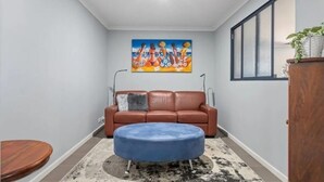A living room with a brown leather sofa, a blue ottoman, a painting on the wall, and a window. - Warm, Cosy winter getaway with Hot Chocolate and Coffee and Heating every room! (Vasse)