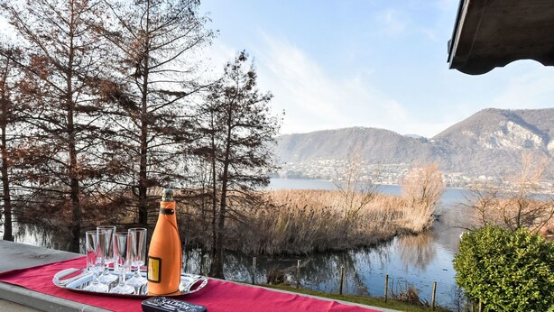 A table with a bottle, glasses, and a remote control on a red mat overlooking a lake and mountains. - La Casetta sul Lago by Interhome (Iseo)