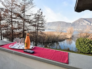 Una mesa con una botella, vasos y un control remoto sobre un tapete rojo, con vistas a un lago y montañas.