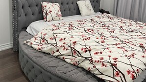 Ein Schlafzimmer mit einem gesteppten, grauen Kopfteil, einer zierlichen Bettdecke mit Blumenmuster und einer grau gepolsterten Bank.