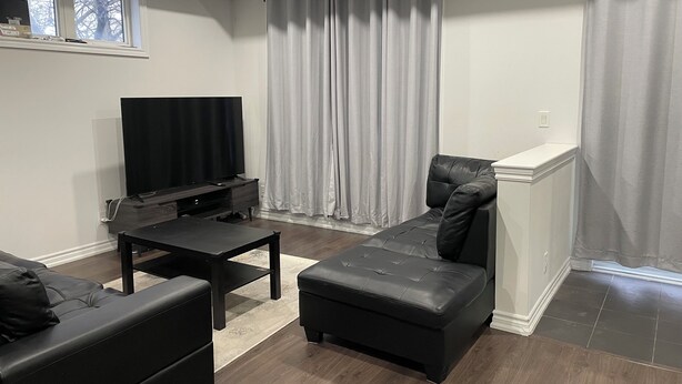 Ein modernes Wohnzimmer mit Flachbildfernseher, schwarzen Ledersofas und einem großen Fenster mit Vorhängen.