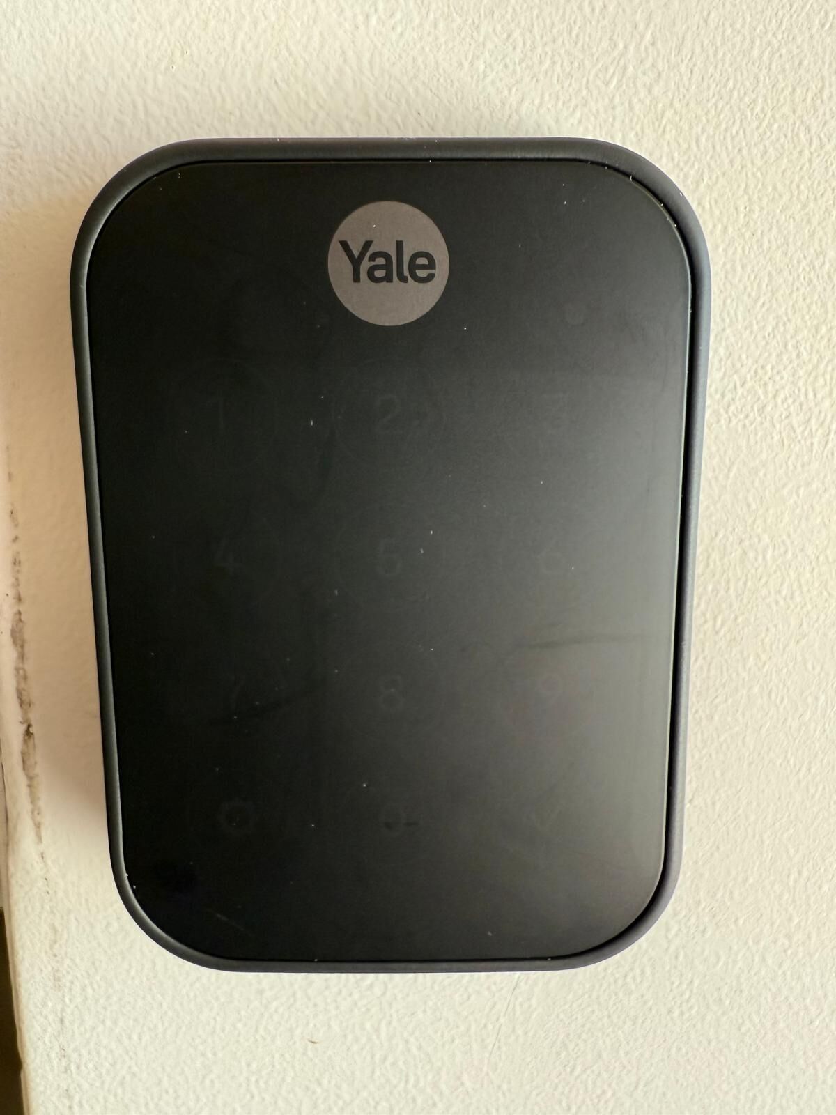 Een zwart Yale apparaat met een cirkelvormig logo op een gestructureerde muur.