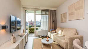 Un salon moderne avec un canapé, une table basse et un téléviseur. Une porte-fenêtre coulissante donne accès à un balcon d’où l’on peut admirer la vue sur les bâtiments et la verdure.