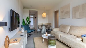 Un salon moderne avec un canapé, une table basse et un fauteuil. On aperçoit une table à manger avec des chaises au fond.