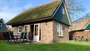 Une maison avec un toit de chaume, des murs en briques et une petite terrasse avec une table et des chaises.