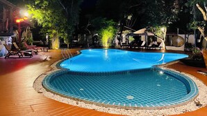 Una piscina illuminata di notte con bordi piastrellati, circondata da poltrone prendisole e alberi.
