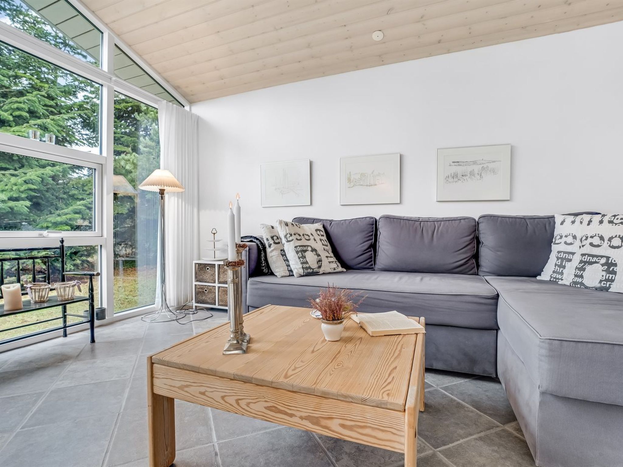 A modern living room with a grey sofa, wooden coffee table, and a large window offering a view of greenery.