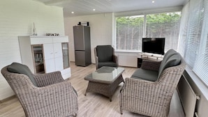 Ein modernes Wohnzimmer mit Rattanmöbeln, einem weißen Schrank, einem grauen Kühlschrank und einem Fernseher in der Nähe eines Fensters mit Jalousien.